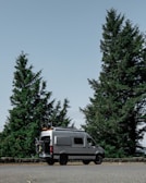 A silver camper van is parked on a roadside with large evergreen trees in the background. The sky is clear, creating a calm and serene atmosphere. The van has equipment attached to its roof, suggesting it is prepared for travel or adventure.