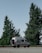 A silver camper van is parked on a roadside with large evergreen trees in the background. The sky is clear, creating a calm and serene atmosphere. The van has equipment attached to its roof, suggesting it is prepared for travel or adventure.