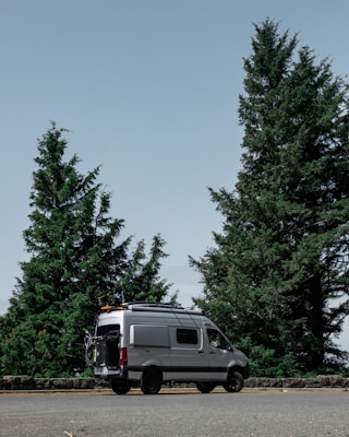 A silver camper van is parked on a roadside with large evergreen trees in the background. The sky is clear, creating a calm and serene atmosphere. The van has equipment attached to its roof, suggesting it is prepared for travel or adventure.