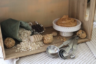 A cozy kitchen scene featuring a wooden box filled with baking ingredients. Rolled oats spill from a green fabric bag, surrounded by a wooden honey dipper and a few energy balls. A small white dish holds creamy peanut butter. Measuring spoons and foil-wrapped chocolate chips are nearby on a textured surface.