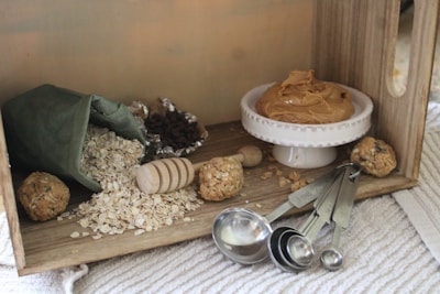 A cozy kitchen scene featuring a wooden box filled with baking ingredients. Rolled oats spill from a green fabric bag, surrounded by a wooden honey dipper and a few energy balls. A small white dish holds creamy peanut butter. Measuring spoons and foil-wrapped chocolate chips are nearby on a textured surface.