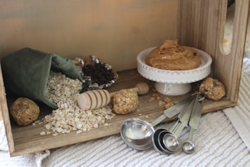 A cozy kitchen scene featuring a wooden box filled with baking ingredients. Rolled oats spill from a green fabric bag, surrounded by a wooden honey dipper and a few energy balls. A small white dish holds creamy peanut butter. Measuring spoons and foil-wrapped chocolate chips are nearby on a textured surface.