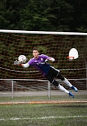 A soccer goalkeeper in a purple jersey is making a diving save in front of a net. The goalkeeper is fully airborne, with arms outstretched toward a soccer ball, wearing gloves and cleats. The netting is orange, and trees are visible in the background.