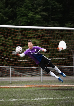 A soccer goalkeeper in a purple jersey is making a diving save in front of a net. The goalkeeper is fully airborne, with arms outstretched toward a soccer ball, wearing gloves and cleats. The netting is orange, and trees are visible in the background.