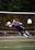 A soccer goalkeeper in a purple jersey is making a diving save in front of a net. The goalkeeper is fully airborne, with arms outstretched toward a soccer ball, wearing gloves and cleats. The netting is orange, and trees are visible in the background.