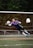 A soccer goalkeeper in a purple jersey is making a diving save in front of a net. The goalkeeper is fully airborne, with arms outstretched toward a soccer ball, wearing gloves and cleats. The netting is orange, and trees are visible in the background.