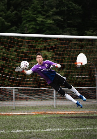 A soccer goalkeeper in a purple jersey is making a diving save in front of a net. The goalkeeper is fully airborne, with arms outstretched toward a soccer ball, wearing gloves and cleats. The netting is orange, and trees are visible in the background.
