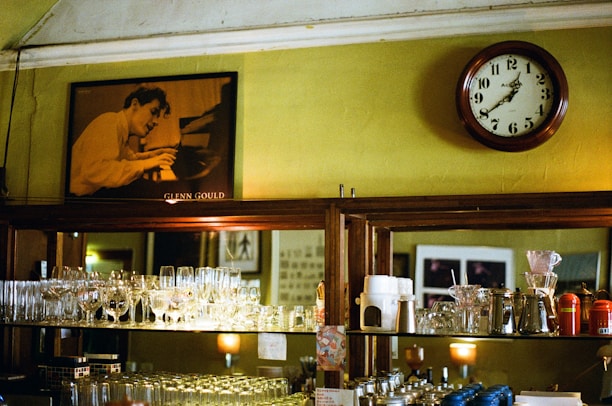 A vintage-style interior with wooden shelves holding an assortment of glassware and coffee equipment. A large round clock with classic numerals hangs on a mustard-colored wall. Above the shelving unit is a black-and-white portrait of a person intensely playing a piano. The setting has a warm and nostalgic feel, reminiscent of an old café or bar.