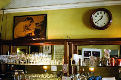 A vintage-style interior with wooden shelves holding an assortment of glassware and coffee equipment. A large round clock with classic numerals hangs on a mustard-colored wall. Above the shelving unit is a black-and-white portrait of a person intensely playing a piano. The setting has a warm and nostalgic feel, reminiscent of an old caf&eacute; or bar.