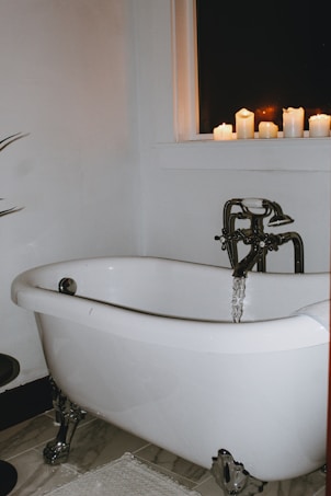 A white clawfoot bathtub is partially filled with running water. Several lit candles are placed on the windowsill, casting a warm glow. The scene is set against a dark window, with a minimalist and cozy atmosphere. A plant is partially visible to the left.