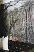 A mural depicting a lush rainforest scene with tall palm trees and dense foliage covers a wall. The corner of a dark leather sofa with tufted detailing is visible in the foreground, along with a couple of white pillows.