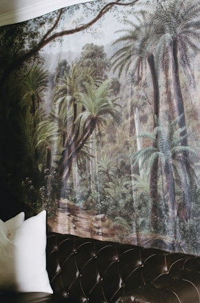 A mural depicting a lush rainforest scene with tall palm trees and dense foliage covers a wall. The corner of a dark leather sofa with tufted detailing is visible in the foreground, along with a couple of white pillows.