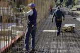 Construction workers wearing protective clothing and gear are working on a building site. They are engaged in assembling metal rebar structures, which are part of the framework for reinforced concrete. The site appears to be set in a mountainous, natural environment.