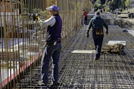 Construction workers wearing protective clothing and gear are working on a building site. They are engaged in assembling metal rebar structures, which are part of the framework for reinforced concrete. The site appears to be set in a mountainous, natural environment.