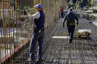 Construction workers wearing protective clothing and gear are working on a building site. They are engaged in assembling metal rebar structures, which are part of the framework for reinforced concrete. The site appears to be set in a mountainous, natural environment.