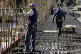 Construction workers wearing protective clothing and gear are working on a building site. They are engaged in assembling metal rebar structures, which are part of the framework for reinforced concrete. The site appears to be set in a mountainous, natural environment.
