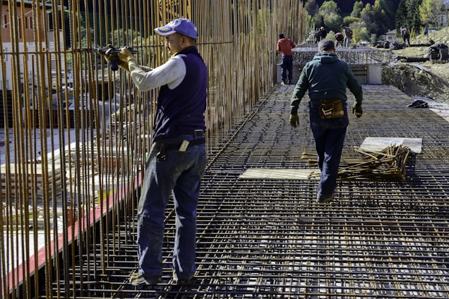 Construction workers wearing protective clothing and gear are working on a building site. They are engaged in assembling metal rebar structures, which are part of the framework for reinforced concrete. The site appears to be set in a mountainous, natural environment.