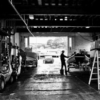 A person stands in a large industrial space, appearing to work on a boat situated inside a warehouse. Several other boats and machinery, including a forklift, are parked along the sides. Large openings allow natural light to illuminate parts of the interior, and a road or courtyard can be seen through one of the openings. Trees and buildings are visible in the background.