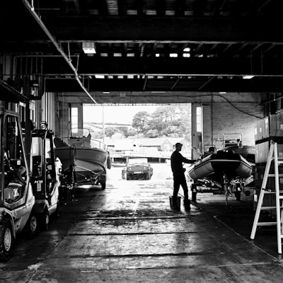 A person stands in a large industrial space, appearing to work on a boat situated inside a warehouse. Several other boats and machinery, including a forklift, are parked along the sides. Large openings allow natural light to illuminate parts of the interior, and a road or courtyard can be seen through one of the openings. Trees and buildings are visible in the background.
