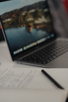 A slightly blurred laptop with a nature-themed desktop wallpaper, on a table next to an open notebook filled with handwritten notes and a pen placed across the pages.