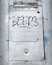 A metal surface with visible wear and peeling paint features a rectangular panel secured by a hinge at the top. The word 'BEERS' is written in a bold, black graffiti style on the central panel.