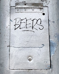 A metal surface with visible wear and peeling paint features a rectangular panel secured by a hinge at the top. The word 'BEERS' is written in a bold, black graffiti style on the central panel.