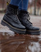 A pair of black leather boots with thick soles is shown in close-up. The boots have yellow stitching and are being worn by someone in blue jeans. They are standing on a wet surface with a reflection visible in a puddle.