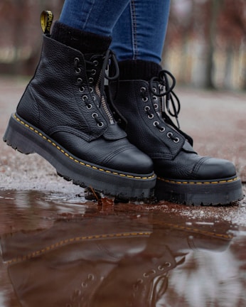 A pair of black leather boots with thick soles is shown in close-up. The boots have yellow stitching and are being worn by someone in blue jeans. They are standing on a wet surface with a reflection visible in a puddle.