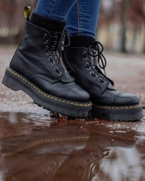 A pair of black leather boots with thick soles is shown in close-up. The boots have yellow stitching and are being worn by someone in blue jeans. They are standing on a wet surface with a reflection visible in a puddle.