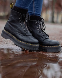 A pair of black leather boots with thick soles is shown in close-up. The boots have yellow stitching and are being worn by someone in blue jeans. They are standing on a wet surface with a reflection visible in a puddle.