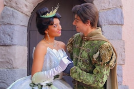 A couple in elaborate, fantasy-themed clothing stands in front of a stone wall. The woman wears a shimmering gown with embellishments, a tiara, and gloves, while the man is in a green, detailed tunic with intricate patterns. They gaze at each other affectionately.