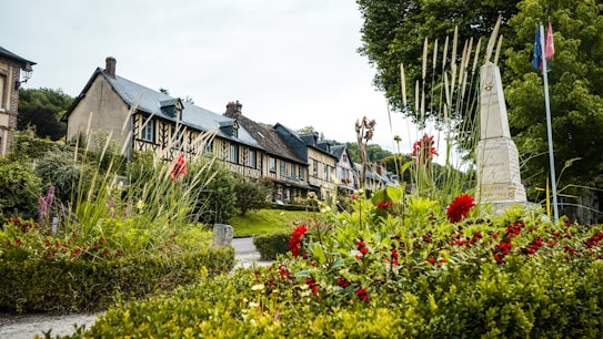 A picturesque, quaint village street with half-timbered houses lined along a narrow road. A lush garden with vibrant red and pink flowers is in the foreground, alongside a stone monument. Tall trees and well-maintained hedges frame the scene, creating a serene and peaceful rural atmosphere.