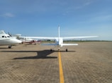 A small white single-engine aircraft is parked on an airfield with clear blue skies overhead. Surrounding it are several other small aircraft positioned on the paved runway, with some in the background. The airfield is expansive, bordered by a green grassy area.