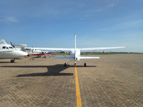 A small white single-engine aircraft is parked on an airfield with clear blue skies overhead. Surrounding it are several other small aircraft positioned on the paved runway, with some in the background. The airfield is expansive, bordered by a green grassy area.