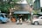 A street scene featuring a small, rustic workshop with an open front, showcasing various tools and machinery inside. Two motorbikes are parked in front, while a blue truck is parked to the left and a beige SUV to the right. The area is shaded by a large tree with lush green leaves, and two ornamental street lamps flank the entrance. The faded, weathered signage above the shop suggests a quaint, local business.