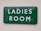 A green rectangular sign with white text reading 'LADIES ROOM' mounted on an off-white wall. The sign appears slightly reflective and has some minor imperfections or dents along the edges.
