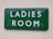 A green rectangular sign with white text reading 'LADIES ROOM' mounted on an off-white wall. The sign appears slightly reflective and has some minor imperfections or dents along the edges.