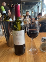 A bottle of Vecchio Ceppo Primitivo Salento wine is positioned prominently on a wooden table. Next to it is a glass partially filled with red wine. An additional bottle of wine, closed, is placed in an ice bucket nearby. In the background, a few people are seated at tables, suggesting a casual dining environment with ambient lighting.