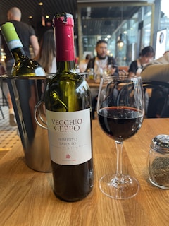 A bottle of Vecchio Ceppo Primitivo Salento wine is positioned prominently on a wooden table. Next to it is a glass partially filled with red wine. An additional bottle of wine, closed, is placed in an ice bucket nearby. In the background, a few people are seated at tables, suggesting a casual dining environment with ambient lighting.