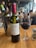 A bottle of Vecchio Ceppo Primitivo Salento wine is positioned prominently on a wooden table. Next to it is a glass partially filled with red wine. An additional bottle of wine, closed, is placed in an ice bucket nearby. In the background, a few people are seated at tables, suggesting a casual dining environment with ambient lighting.