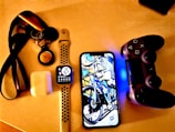 A collection of personal electronic items and accessories on a light-colored surface, including a smart watch with a black and white strap displaying the time, a smartphone showing an image of a bicycle, a game controller with a glowing blue light, a pair of wireless earbuds in a charging case, and a set of keys attached to a lanyard.
