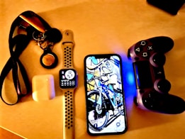 A collection of personal electronic items and accessories on a light-colored surface, including a smart watch with a black and white strap displaying the time, a smartphone showing an image of a bicycle, a game controller with a glowing blue light, a pair of wireless earbuds in a charging case, and a set of keys attached to a lanyard.