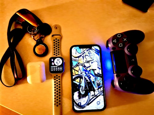A collection of personal electronic items and accessories on a light-colored surface, including a smart watch with a black and white strap displaying the time, a smartphone showing an image of a bicycle, a game controller with a glowing blue light, a pair of wireless earbuds in a charging case, and a set of keys attached to a lanyard.