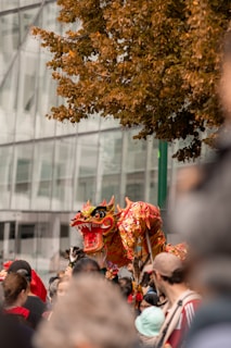 A vibrant dragon puppet is held above a large crowd, likely at a festival or parade. The dragon is intricately designed with red, gold, and black colors, showing a fierce expression. The background features a modern glass building and a tree with brown leaves.