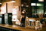 A person is preparing a hot beverage behind a counter in a cozy cafe setting. Various coffee-making equipment and supplies are neatly organized, with shelves holding mugs and a background featuring dim lighting.