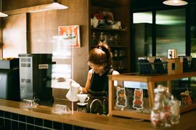 A person is preparing a hot beverage behind a counter in a cozy cafe setting. Various coffee-making equipment and supplies are neatly organized, with shelves holding mugs and a background featuring dim lighting.