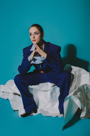 A person dressed in a dark blue suit is seated on crumpled white papers against a teal background. The individual has a thoughtful expression, with hands clasped near their face. They exhibit an introspective mood with slicked-back hair and dramatic eye makeup.