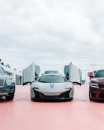 Three luxury cars are parked side by side in an open area with overcast skies above. The central car, a sleek sports car, has its doors open upwards, revealing its futuristic design. On either side, a large SUV and another car with a muscular build complete the lineup.