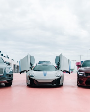 Three luxury cars are parked side by side in an open area with overcast skies above. The central car, a sleek sports car, has its doors open upwards, revealing its futuristic design. On either side, a large SUV and another car with a muscular build complete the lineup.