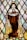 A stained glass artwork depicting a religious figure dressed in a brown robe and white cloak, holding a banner that reads 'Follow Him.' The figure has a halo and three red roses on the robe. The background consists of diamond-shaped pieces of glass in subtle colors.